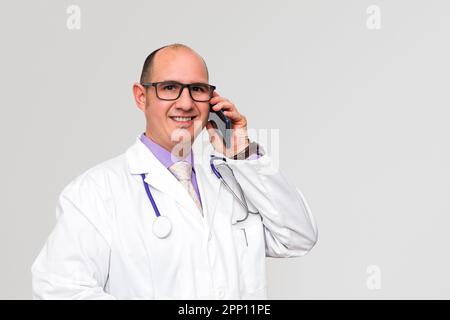 Ein glatzköpfiger, lächelnder, weißer Arzt mit Brille, der einen weißen Mantel über einem lila Hemd trägt, führt ein Gespräch mit seinem Handy. Backgro Stockfoto