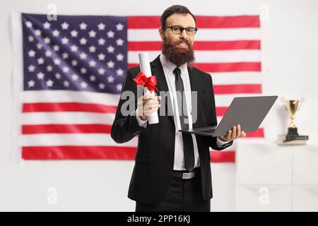 Bärtiger Mann in einem Anzug mit einem Abschlusszeugnis und einem Laptop vor der US-Flagge Stockfoto
