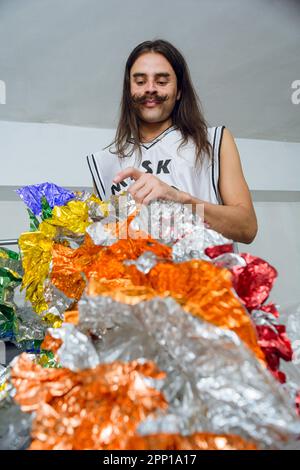 Porträt eines nicht-binären lateinamerikanischen Mannes in seinem Zimmer, der sein farbenfrohes Drag-Queen-Kostüm aus recyceltem Material herstellt Stockfoto