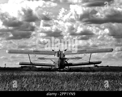 Isoliertes altes Doppeldecker-Flugzeug auf einem dunklen Wiesen-Flugplatz unter dynamischem Himmel mit weißen Wolken von hinten. Schwarzweißbild. Segelflugzeug. Stockfoto