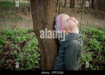 Kleinmädchenpraxen. Das Mädchen lehnte sich mit dem Rücken gegen den Baum und schaute hoch, genoss die Energie der Natur. Baumtherapie Stockfoto