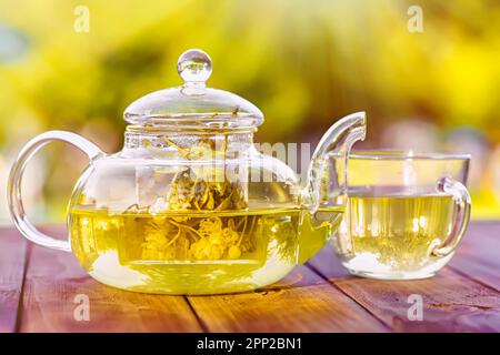 Teekanne gefüllt mit heißem Lindentee und Lindenblumen, auf einem Holztisch im Garten. Die rustikale Atmosphäre trägt zum Charme des nicht traditionellen Kräutertees bei Stockfoto