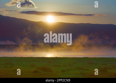 Grünes Glasfeld, See und Berge im Morgennebel. Stockfoto