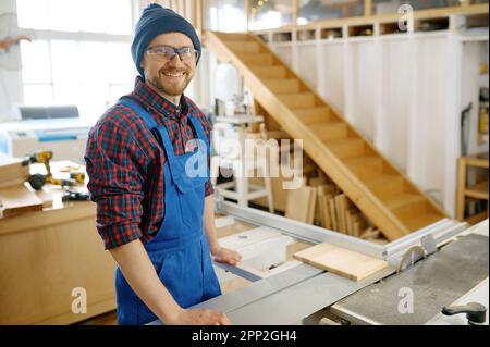 Porträt eines fröhlichen lächelnden Zimmermanns im Overall, der am Arbeitsplatz steht Stockfoto