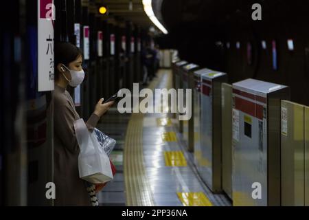 Eine junge japanische Frau, die eine chirurgische Maske trägt, benutzt ihr Smartphone, während sie auf einen Zug am Bahnhof Roppongi mit Bahnsteig-Sicherheitsbarrieren wartet Stockfoto
