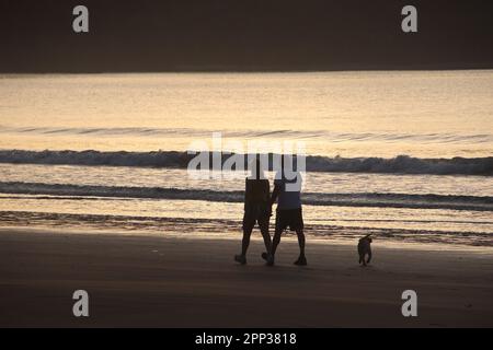 Während die Sonne über den Horizont blickt, spazieren zwei Personen mit ihrem Hundebegleiter am Strand entlang, das sanfte Wellenschlagen sorgt für einen beruhigenden Soun Stockfoto