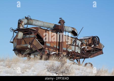 Eine verlassene antike Dreschmaschine ähnelt einer Metallechse auf einem schneebedeckten Hang auf der Ebene von Montana Stockfoto