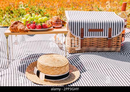 Sommer-Picknick-Komposition, Korb mit Korb, Strohhut und kleiner Tisch mit Obst und Croissants auf Blumenwiesen Stockfoto