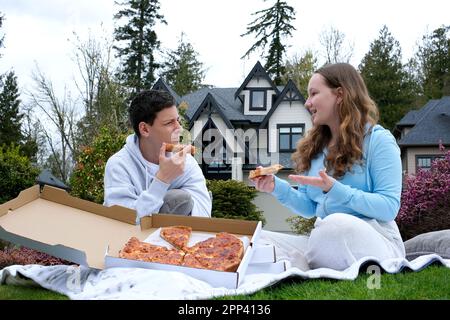 Teenager, Junge und Mädchen, die sich unterhalten und Pizza essen. Sie sitzen in der Natur in der Nähe des Hauses. Teenager, die sich in frischer Luft entspannen. Die ersten Sonnenstrahlen im Frühling gehen raus, sitzen auf dem Gras und kommunizieren offline Stockfoto