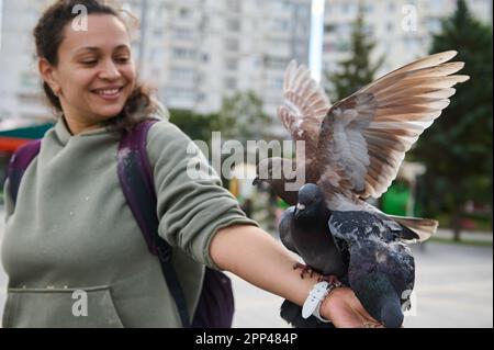 Lächelnde afrikanische Amrikanerin, positive Frau, die Felsentauben füttert, die auf ihrem ausgestreckten Arm auf dem Stadtplatz sitzt. Konzentrieren Sie sich auf Tauben. Stockfoto