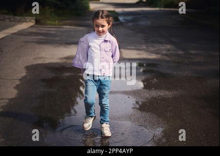 Das ganze Porträt eines wunderschönen, fröhlichen kleinen Mädchens in Jeans und lila Jacke, lächelnd und amüsant, springt auf eine kleine Pfütze Stockfoto