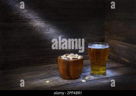 Eine braune, hölzerne Schüssel voller ungesalzener Nüsse und ein Glas Bier in rustikalem Holzambiente in Pools mit stimmungsvoller Beleuchtung; Kopierbereich links Stockfoto