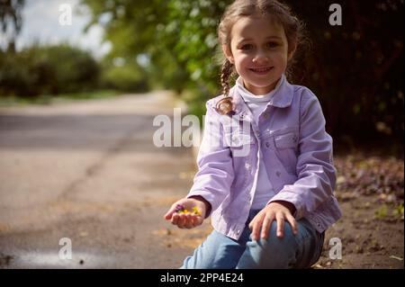 Selbstbewusstes Porträt eines kaukasischen, bezaubernden, positiven Kindes in einer stylischen, lila Jeans und Jacke, die auf Pflastersteinen sitzt, lächelt und süsst Stockfoto