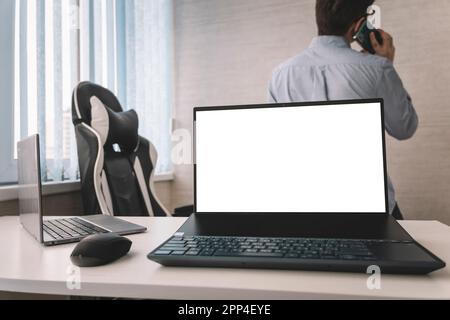 Laptop am Arbeitsplatz in einem weißen Büro vor dem Hintergrund eines Mitarbeiters, der am Telefon spricht. Stockfoto