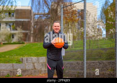 Ein Mann mittleren Alters mit schwarzen Sporthosen hält den Basketball in den Händen und lächelt freundlich vor der Kamera auf einem Sportplatz Stockfoto