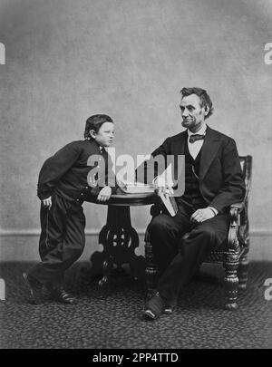 Abraham Lincoln setzte sich und hielt ein Buch mit seinem Sohn Tad (Thomas), der sich auf einen Tisch lehnte. 5. Februar 1865 Alexander Gardner Stockfoto