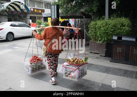 Eine Chinesin trägt Feigen und Rambutane zum Verkauf auf Körben, die an einem Stock auf ihren Schultern hängen - Chengdu, China. Stockfoto