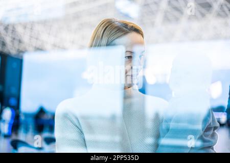 Die nachdenkliche junge Mutter schaute durch das Fenster und hielt sein kleines Kind fest, während sie darauf wartete, an den Abflugsteigen des Flughafenterminals an Bord eines Flugzeugs zu gehen. Das „Reise mit Baby“-Konzept Stockfoto