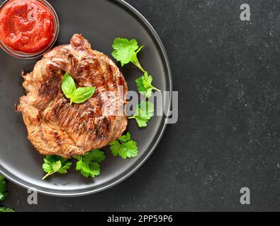 Saftiges gegrilltes Rindersteak und Tomatensauce auf dem Teller auf schwarzem Steinhintergrund mit Kopierbereich. Draufsicht, flach liegend Stockfoto