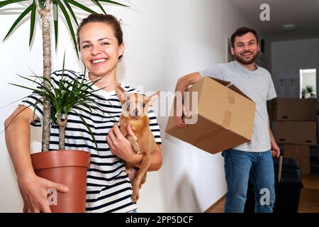 Glückliche junge Familie zieht um und trägt ihren kleinen Hund, ihre Pflanze, Pappschachtel. Stockfoto