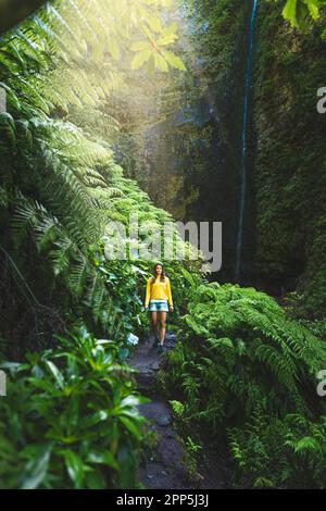 Beschreibung: Touristenfrau, die auf einem mit Farn überwucherten Pfad am malerischen, überwucherten Wasserfall im Madeiras Regenwald spaziert. Levada von Caldeirão Verd Stockfoto