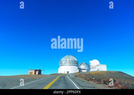 Gepflasterte Straße, die zu den astronomischen Forschungseinrichtungen führt, die sich auf dem Mauna Kea Gipfel vor blauem Himmel befinden. Stockfoto