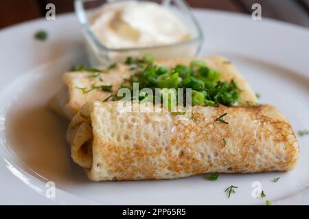 Zwei Hälften Pfannkuchenrolle mit Himbeerfüllung, saure Sahne auf weißem Teller, Teelöffel in Schüssel mit saurer Sahne, Gabel auf Holztisch, selektiver Fokus Stockfoto