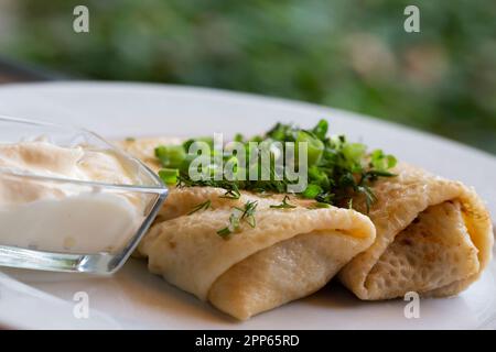 Zwei Hälften Pfannkuchenrolle mit Himbeerfüllung, saure Sahne auf weißem Teller, Teelöffel in Schüssel mit saurer Sahne, Gabel auf Holztisch, selektiver Fokus Stockfoto