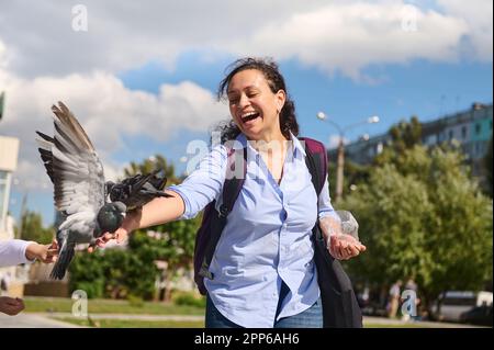 Fröhliche, positiv lächelnde, multiethnische, positive junge Frau, die an einem sonnigen Frühlingstag Tauben auf dem Platz füttert. Das Konzept von Liebe, Fürsorge und Kompassio Stockfoto