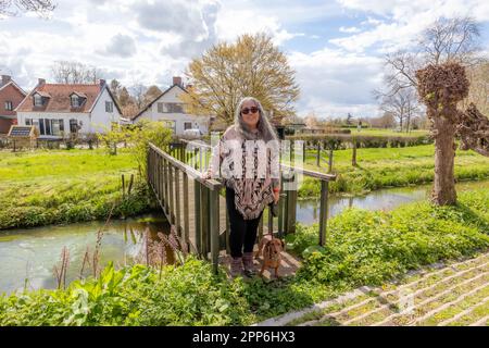 Eine Frau mit ihrem braunen Dackel, die auf einer Brücke über dem Fluss steht, einen indischen Poncho und eine Sonnenbrille trägt, Häuser und Ackerland im Hintergrund gegen BL Stockfoto