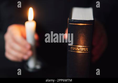 Männerhände mit bibel und zwei brennenden Kerzen während des Gebets auf schwarzem Hintergrund. Die Inschrift auf dem aus Russisch übersetzten Buch: Die Bibel Stockfoto