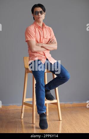 Ein junger Mann mit Brille, sitzt auf einem Stuhl, kreuzt die Arme und schaut in die Kamera Stockfoto