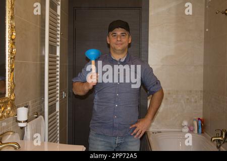 Bild eines lächelnden Klempners im Badezimmer, der einen Stößel in der Hand hält, während er ein Waschbecken aufräumt. Heimwerkerarbeit zu Hause. Stockfoto