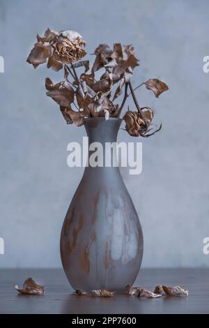 Blumenstrauß aus getrockneten Blumen in Glasvase mit heruntergefallenen Blättern am Boden, in einer kalten gedämpften Farbenatmosphäre vor einem rustikalen Hintergrund. Stockfoto