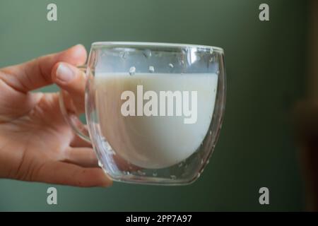 Ein junges ukrainisches Mädchen hält morgens einen durchsichtigen Glasbecher mit Milch Stockfoto