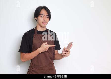 Porträt eines attraktiven asiatischen Barista in brauner Schürze, der ein Mobiltelefon hält und zeigt. Isoliertes Bild auf weißem Hintergrund Stockfoto