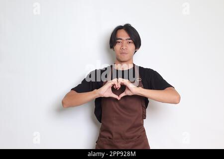 Das Porträt eines attraktiven asiatischen Barista in einer braunen Schürze spricht über eigene Gefühle, macht eine Herzgeste über der Brust, drückt Sympathie und Liebe aus, lächelt Stockfoto