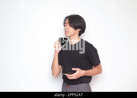 Porträt eines gutaussehenden Asiaten im schwarzen T-Shirt, der eine Tasse Kaffee trinkt, während er die Untertasse hält. Isoliertes Bild auf weißem Hintergrund Stockfoto