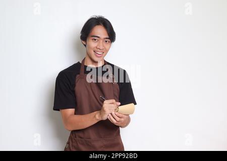 Porträt eines attraktiven asiatischen Barista in brauner Schürze, der auf die Speisekarte schreibt. Isoliertes Bild auf weißem Hintergrund Stockfoto