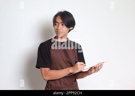 Porträt eines attraktiven asiatischen Barista in brauner Schürze, der auf die Speisekarte schreibt. Isoliertes Bild auf weißem Hintergrund Stockfoto