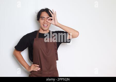 Porträt eines attraktiven asiatischen Baristas in brauner Schürze mit einer guten Handgeste und einem lächelnden Blick in die Kamera. Werbekonzept. Isoliertes Bild ein Stockfoto