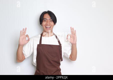 Porträt eines attraktiven asiatischen Baristas in brauner Schürze mit einer guten Handgeste und einem lächelnden Blick in die Kamera. Werbekonzept. Isoliertes Bild ein Stockfoto