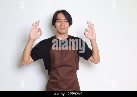 Porträt eines attraktiven asiatischen Baristas in brauner Schürze mit einer guten Handgeste und einem lächelnden Blick in die Kamera. Werbekonzept. Isoliertes Bild ein Stockfoto