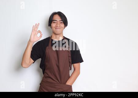 Porträt eines attraktiven asiatischen Baristas in brauner Schürze mit einer guten Handgeste und einem lächelnden Blick in die Kamera. Werbekonzept. Isoliertes Bild ein Stockfoto