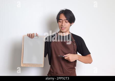 Porträt eines attraktiven asiatischen Baristas in brauner Schürze mit leerem Menüpapier auf dem Klemmbrett. Isoliertes Bild auf weißem Hintergrund Stockfoto
