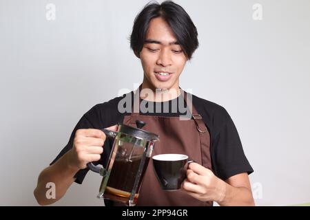 Porträt eines attraktiven asiatischen Baristas in einer braunen Schürze, der Kaffee von der French Press Kaffeemaschine in eine Tasse gießt. Isoliertes Bild auf weißem Hintergrund Stockfoto