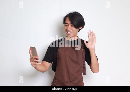 Porträt eines attraktiven asiatischen Baristas in einer braunen Schürze, der während eines Videogesprächs eine gute Geste macht. Isoliertes Bild auf weißem Hintergrund Stockfoto