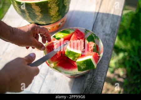 Die Hand eines Wassermelonenmanns auf einem Holztisch zu schneiden. Männerhände beim Schneiden von Wassermelone im Sommer an einem sonnigen Tag. Stockfoto