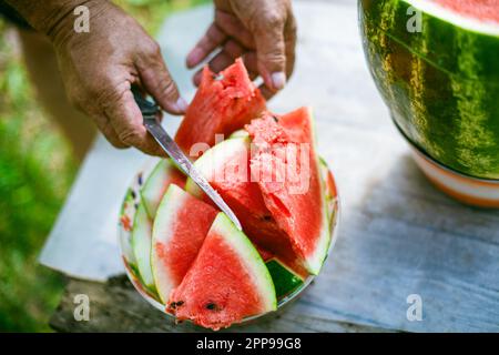 Die Hand eines Wassermelonenmanns auf einem Holztisch zu schneiden. Männerhände beim Schneiden von Wassermelone im Sommer an einem sonnigen Tag. Stockfoto