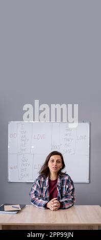 Glückliche Lehrerin sitzt am Holzschreibtisch und schaut vor dem Whiteboard auf die Kamera mit englischen Grammatikregeln im Hintergrund. Lehrerin Stockfoto
